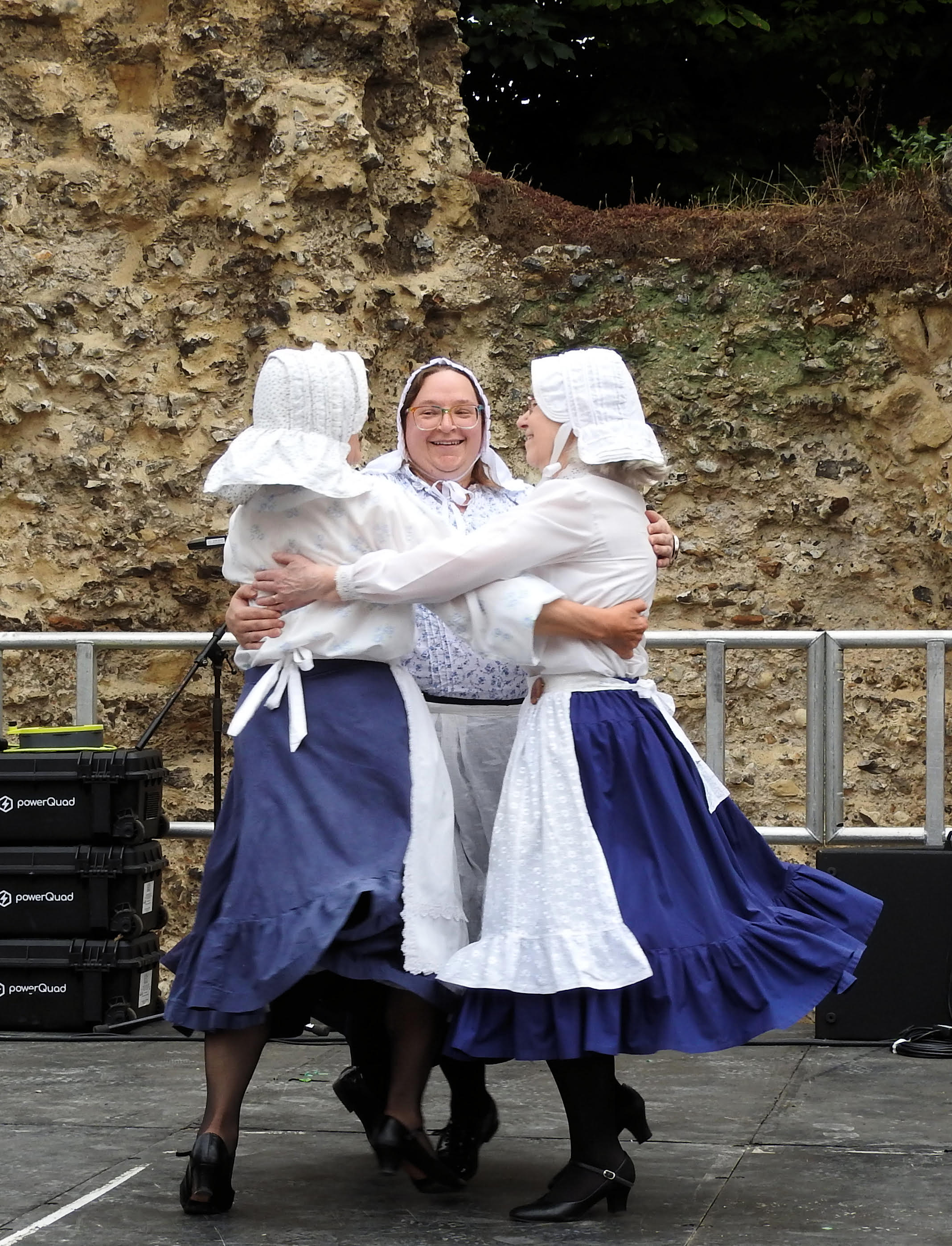Dancers in action. Three women in traditional clothing are shown in a 'basket' dance move, involving close contact. THey are shown in an ourdoor venue.