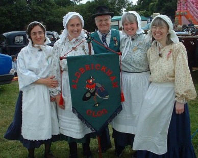 An image of some of Aldbrickham Clog's dancers. Four women, with a man stood in the middle of them, are dressed in traditional 'washerwoman' clothing representing the British industrial era. The women are wearing white aprons and bonnets the man is wearing a bowler hat. They are holding a fabric sign which reads 'Aldbrickham clog' in red writing with a jester-type figure, a black dancing shoe and a clog all entwined together - this is the group's logo. The group are smiling, and are stood outdoors in a field; there are classic cars and a bouncy castle in the background.